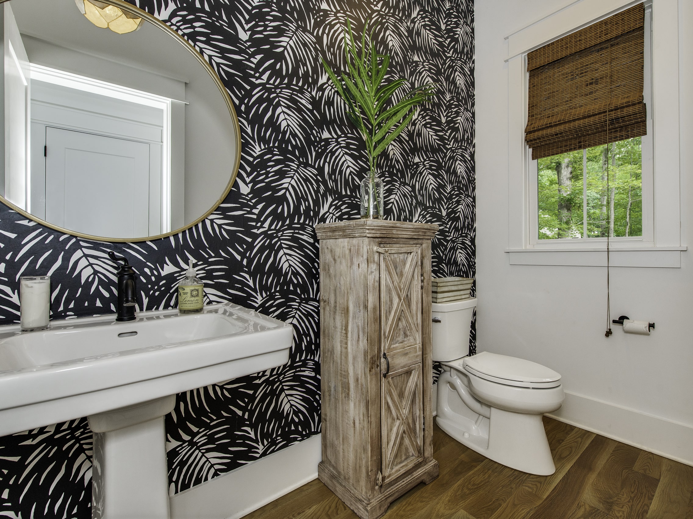Powder room renovation with patterned wallpaper, pedestal sink, wood cabinet, and round mirror
