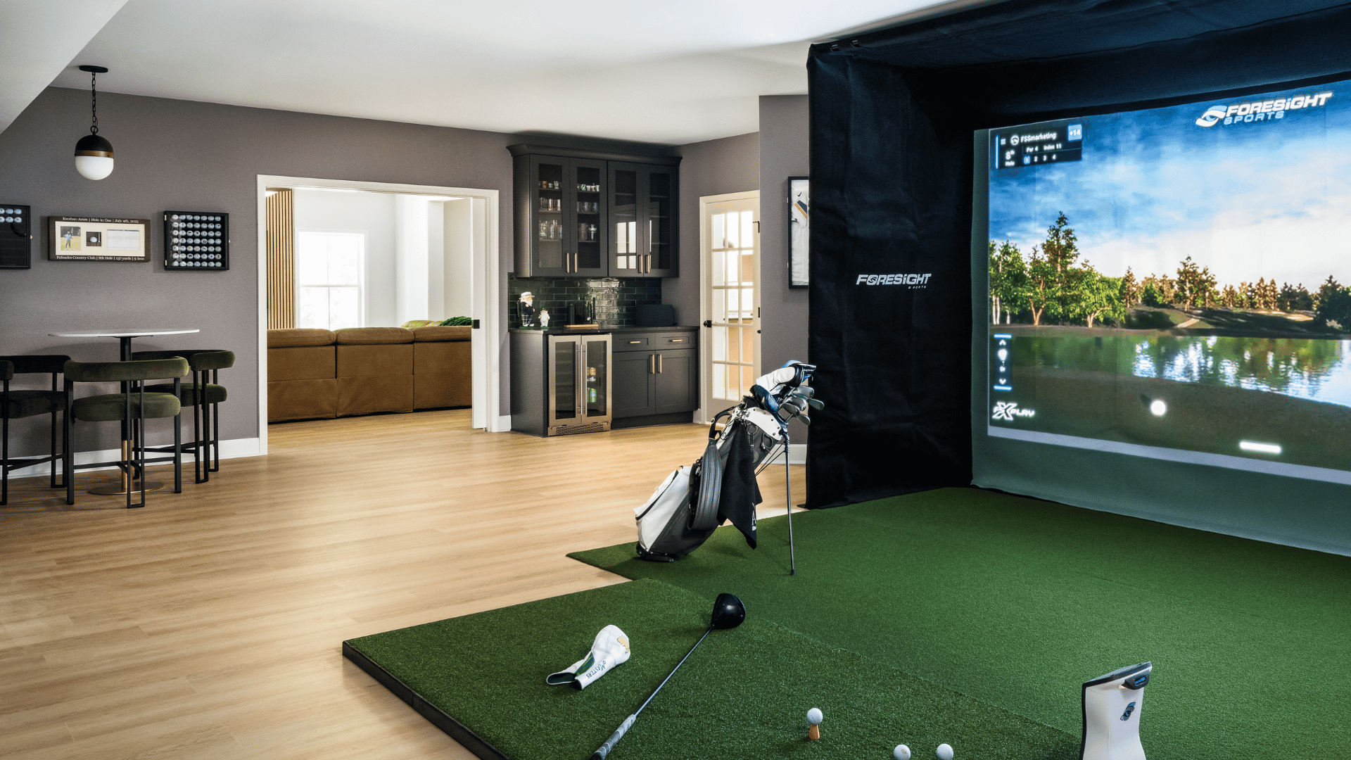 Custom basement golf simulator room with Foresight Sports simulator screen, artificial turf hitting mat, dark gray walls, custom wet bar with wine refrigerator, and lounge seating area built by PKB Design Build in Charlotte NC