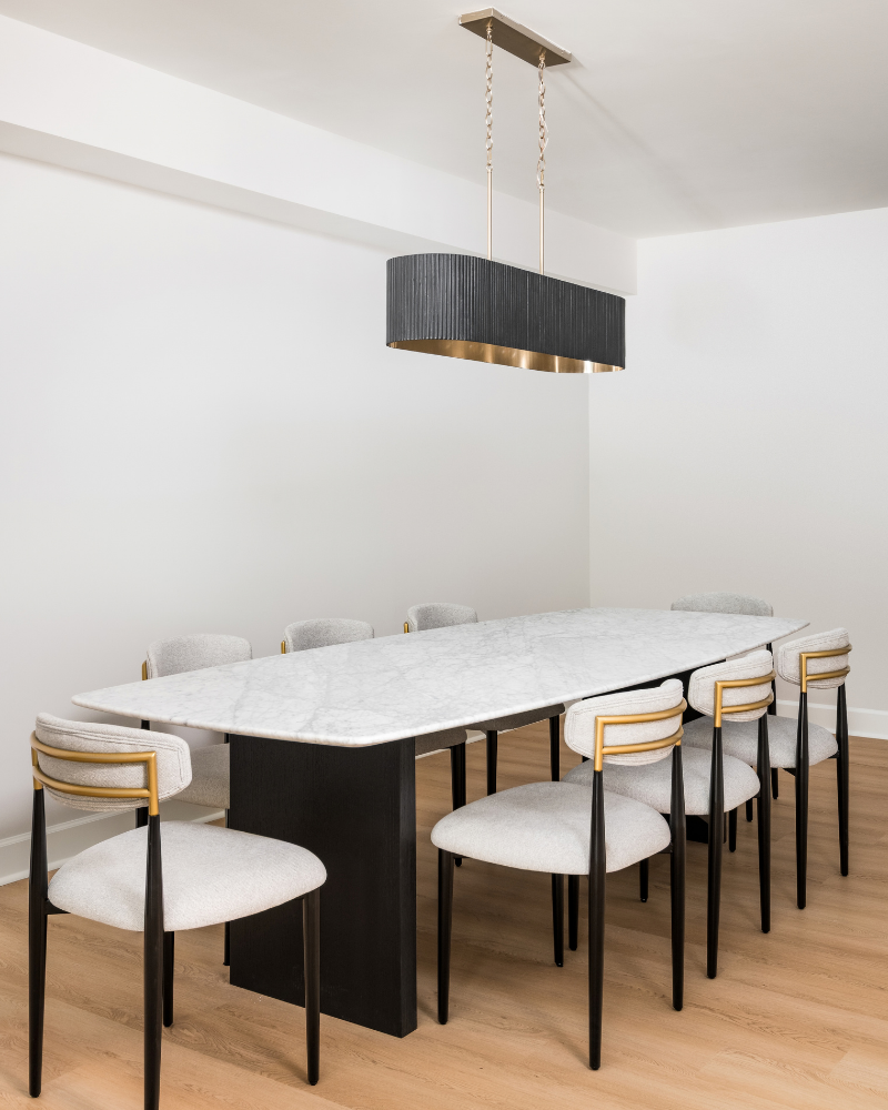 Basement dining area with a white marble table on a black pedestal base, cream upholstered chairs with brass backs, and a black drum chandelier Basement dining area with a white marble table on a black pedestal base, cream upholstered chairs with brass backs, and a black drum chandelier